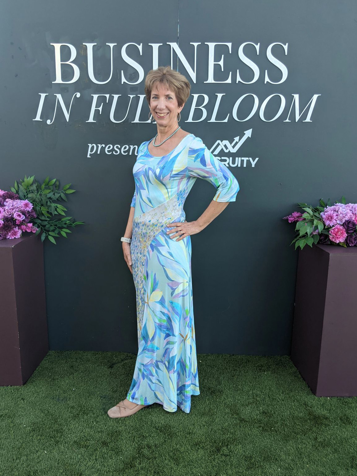 Debbe Garry in formal dress, posing in front of a backdrop reading "Business in Full Bloom"