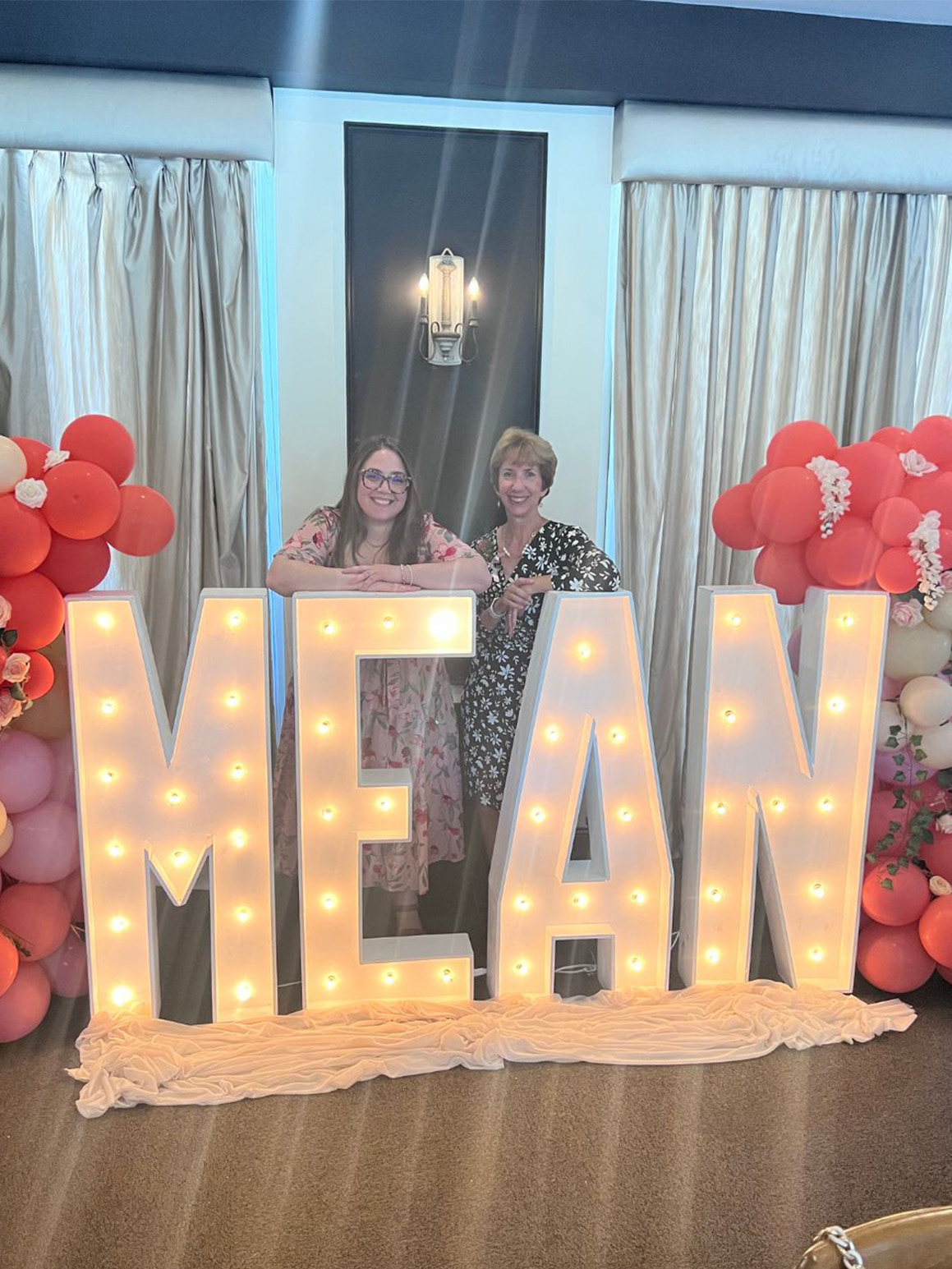 Kayla and Debbe standing behind light up letters spelling "MEAN"